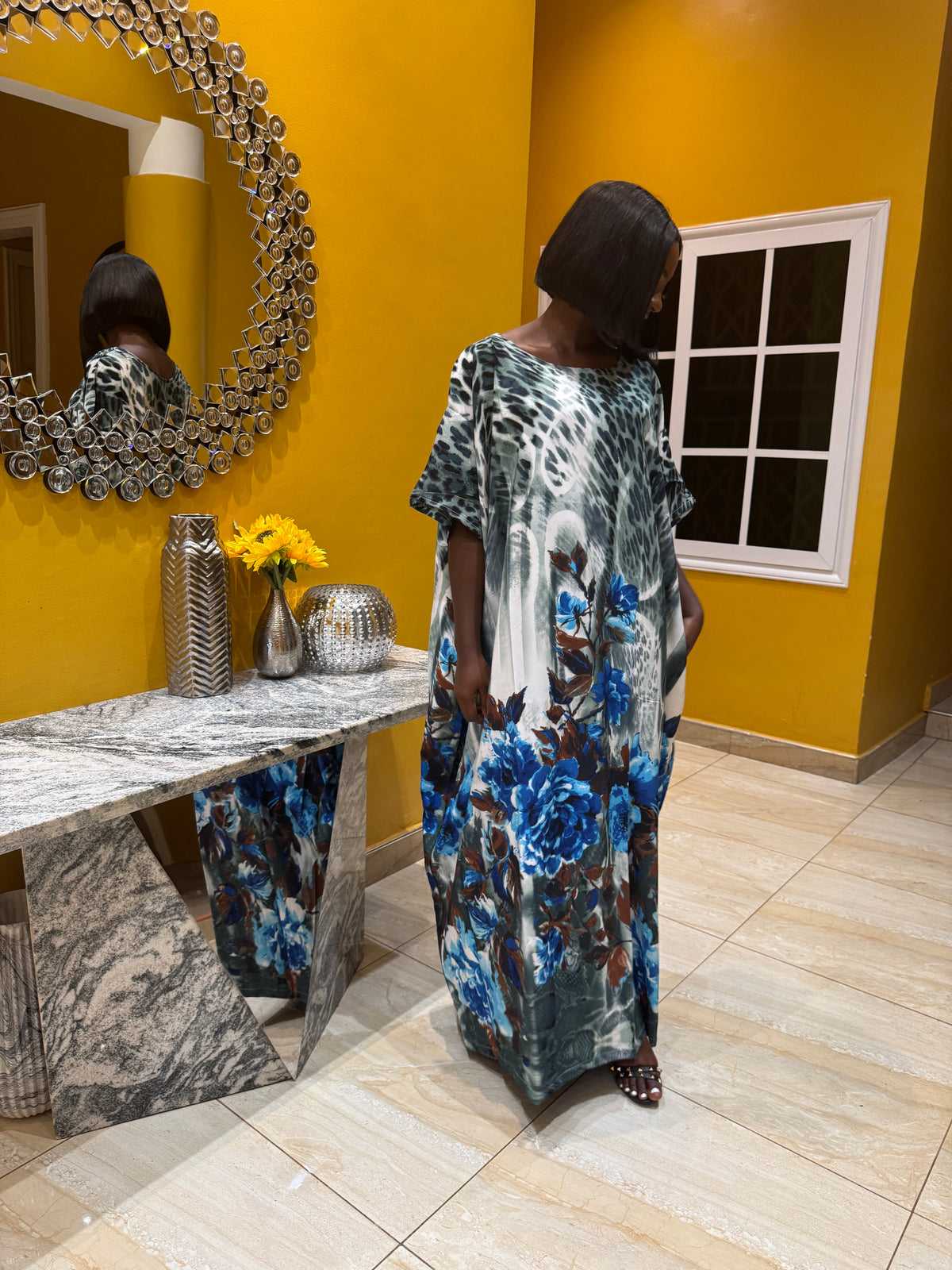 Woman in a floral kaftan dress standing in front of a yellow wall with a mirror.