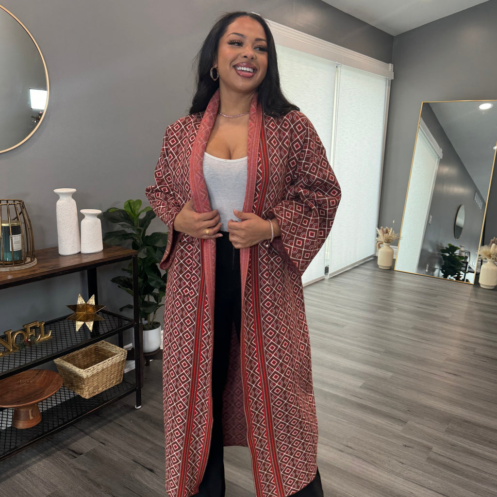 A person wearing a brown knit cardigan with a geometric pattern, standing in a room with light decor.