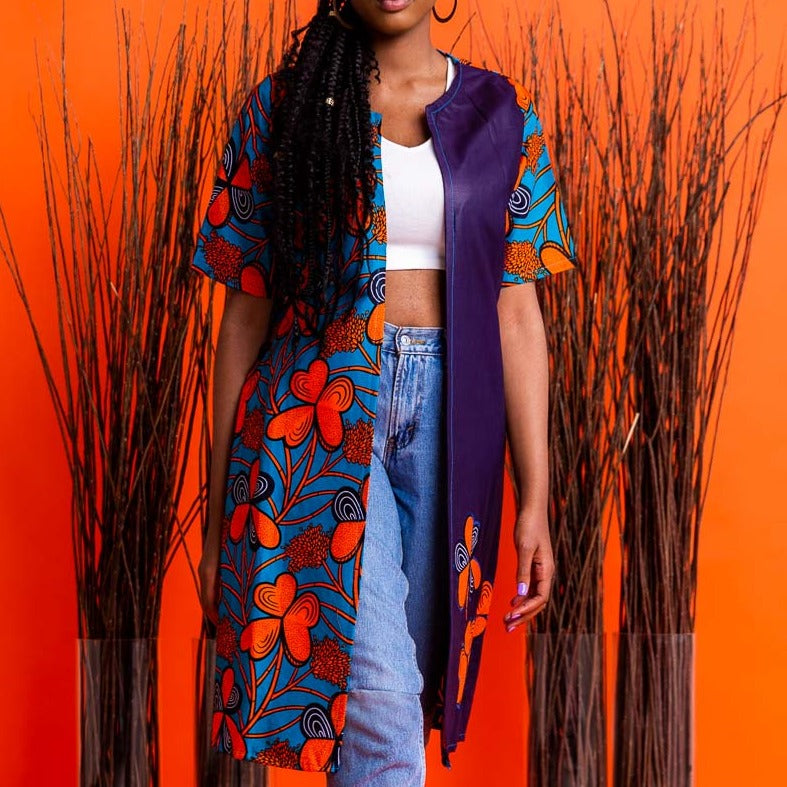 A woman wearing an overcoat with colorful floral African print standing against an orange background with decorative grasses to the side.